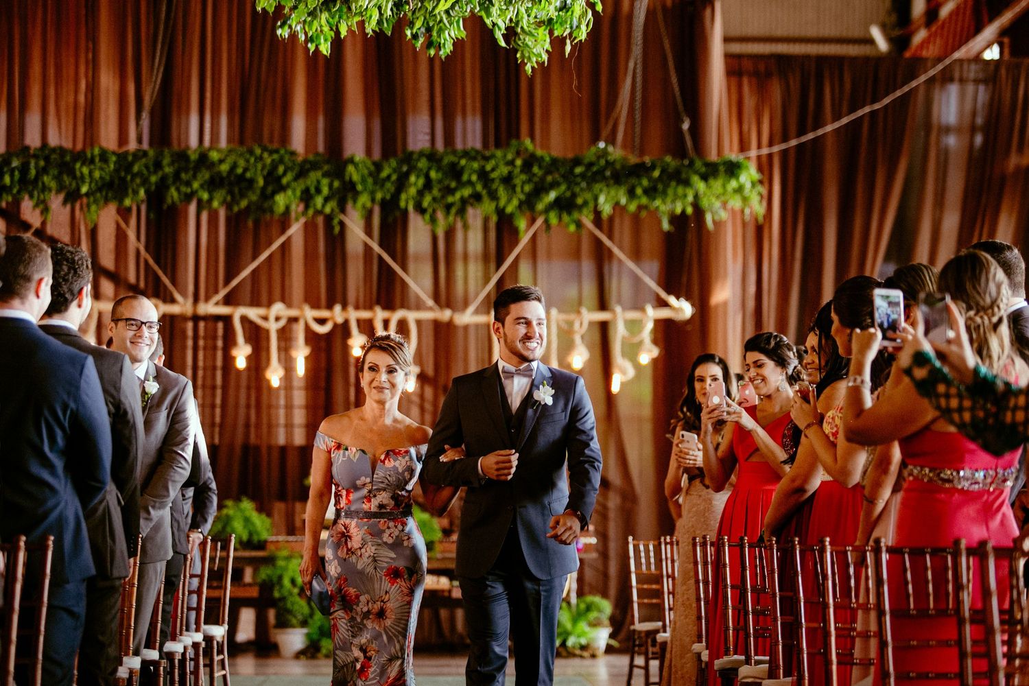 Wedding ceremony in elegant venue with hanging greenery and warm lighting.