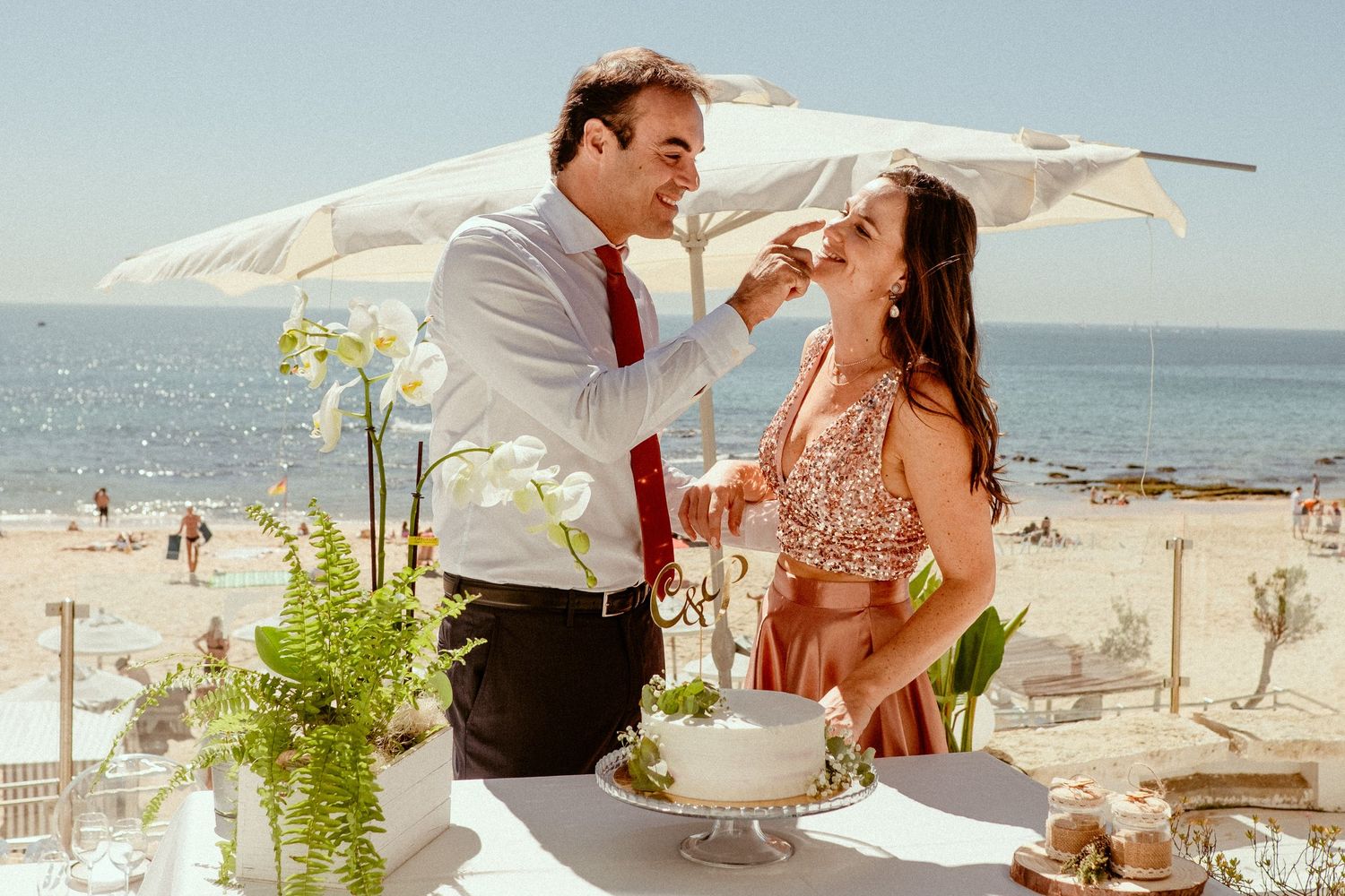 Joyful couple sharing wedding cake on a sunny beach terrace decorated with white orchids and ocean views.