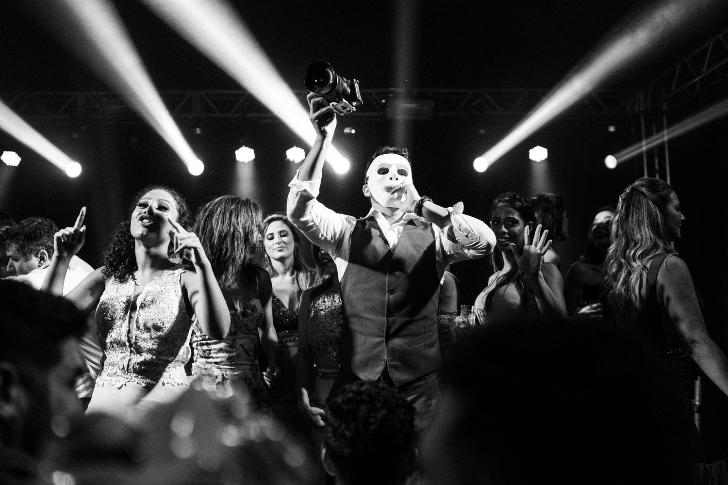 A black and white photo shows celebratory dancing under dramatic spotlights at a wedding reception.