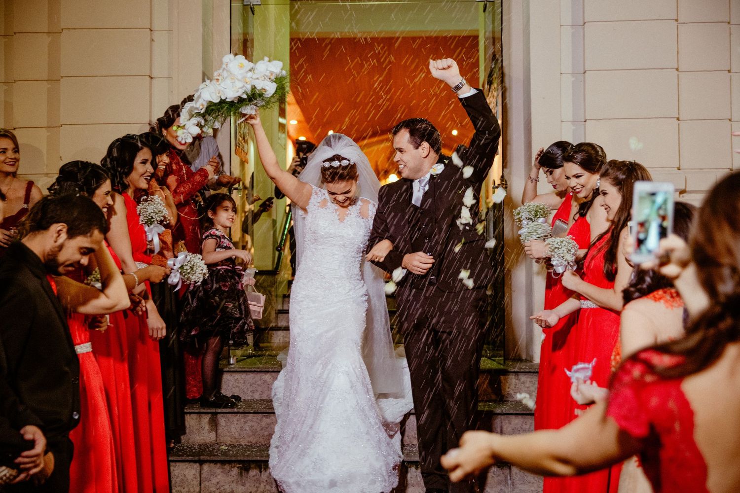 Wedding party in red dresses celebrating outside a white stone building.