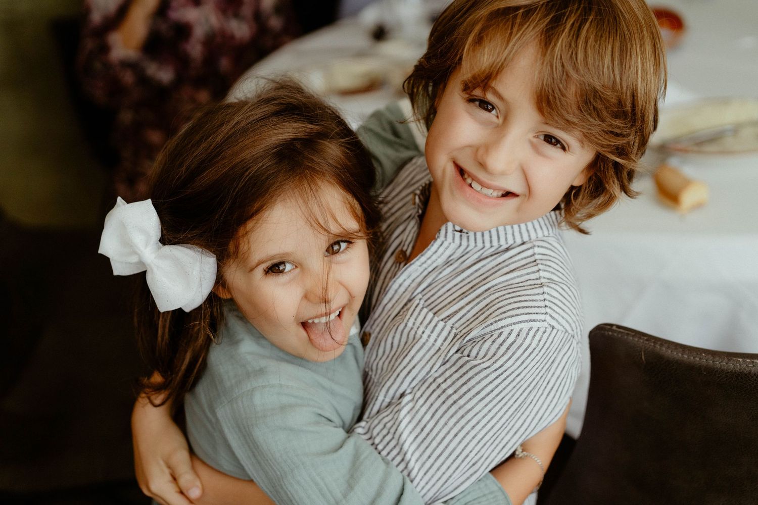 Two children in casual clothes smile and hug while sitting together indoors.