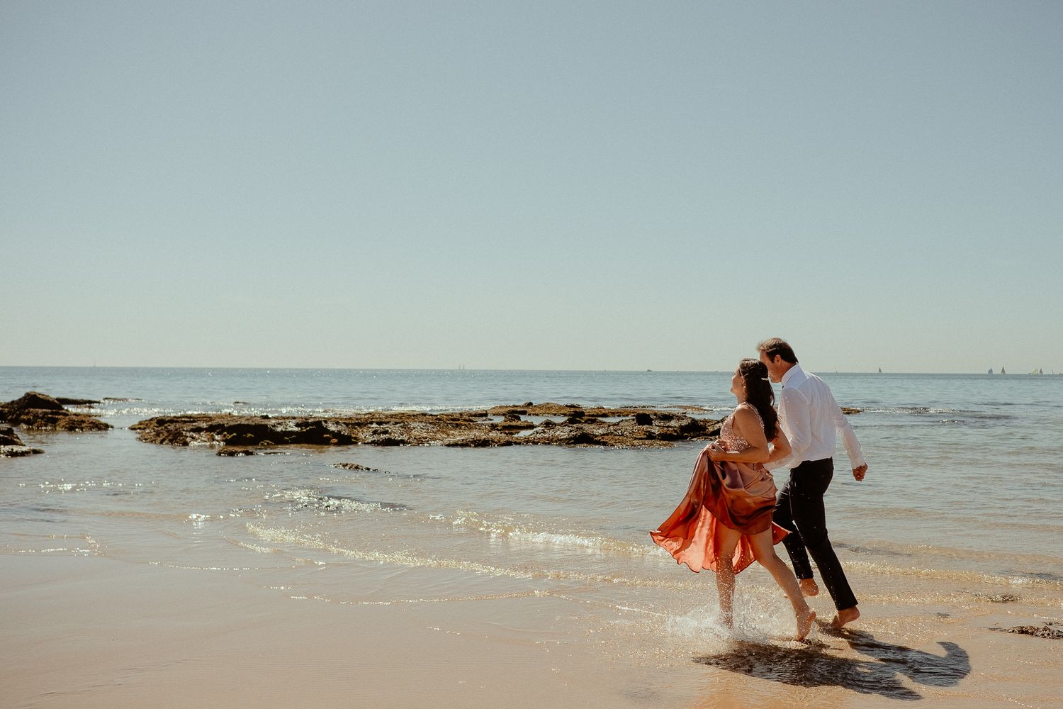 A person in an orange shirt walks along a sandy beach with calm ocean waters and clear blue sky in the background.