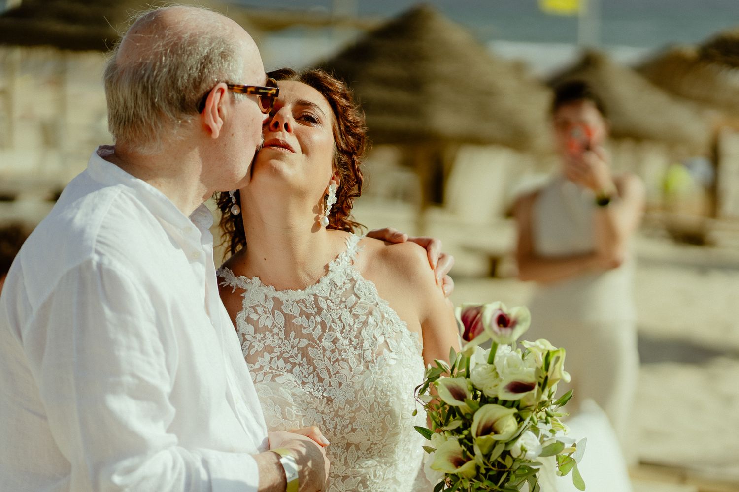 Couple sharing a romantic kiss on a beach wedding, bride holding a green bouquet, officiant standing in background.