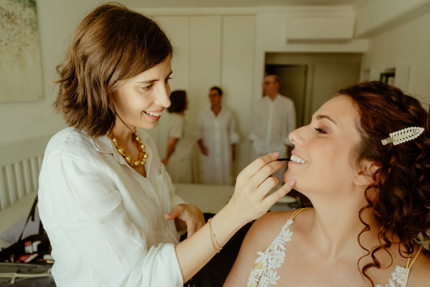 Makeup artist applying finishing touches to a bride in a lace wedding dress before her wedding ceremony.