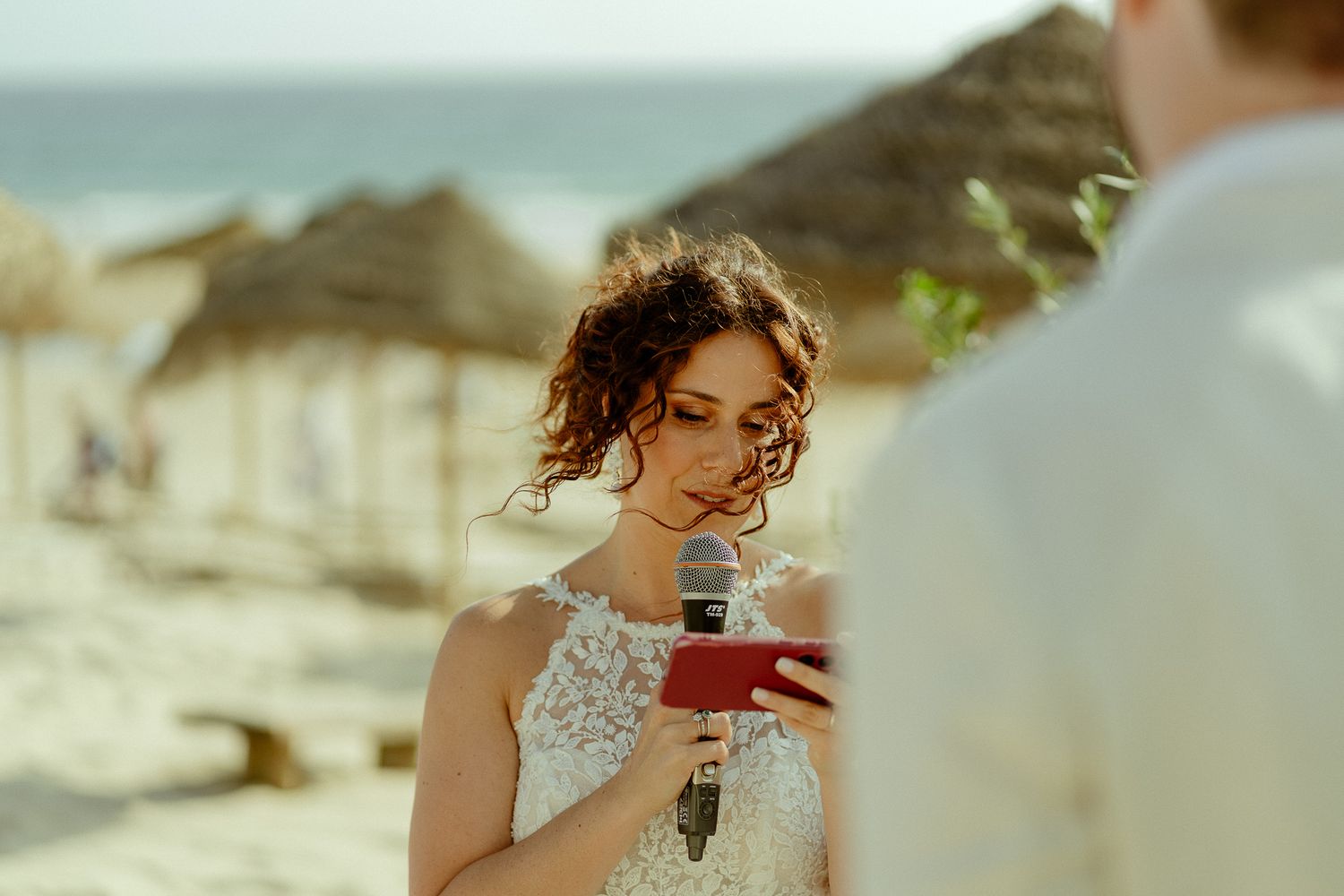 Bride in lace wedding dress reading vows from red phone on a sunny beach with thatched umbrellas in background.