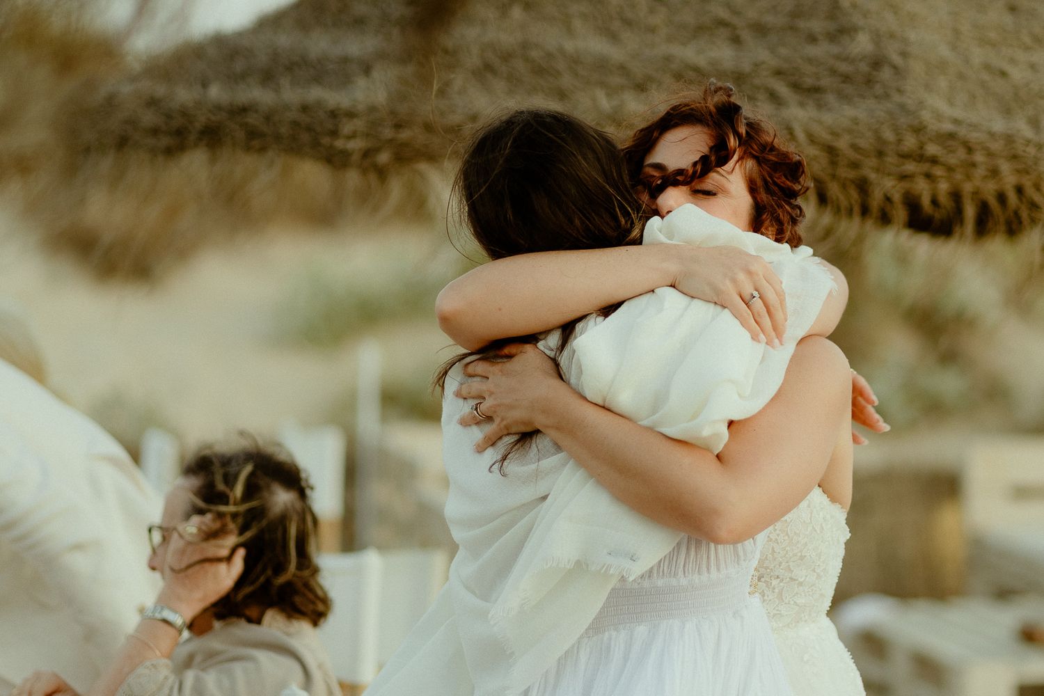 Two women sharing a warm embrace at a beach wedding ceremony in soft neutral tones.