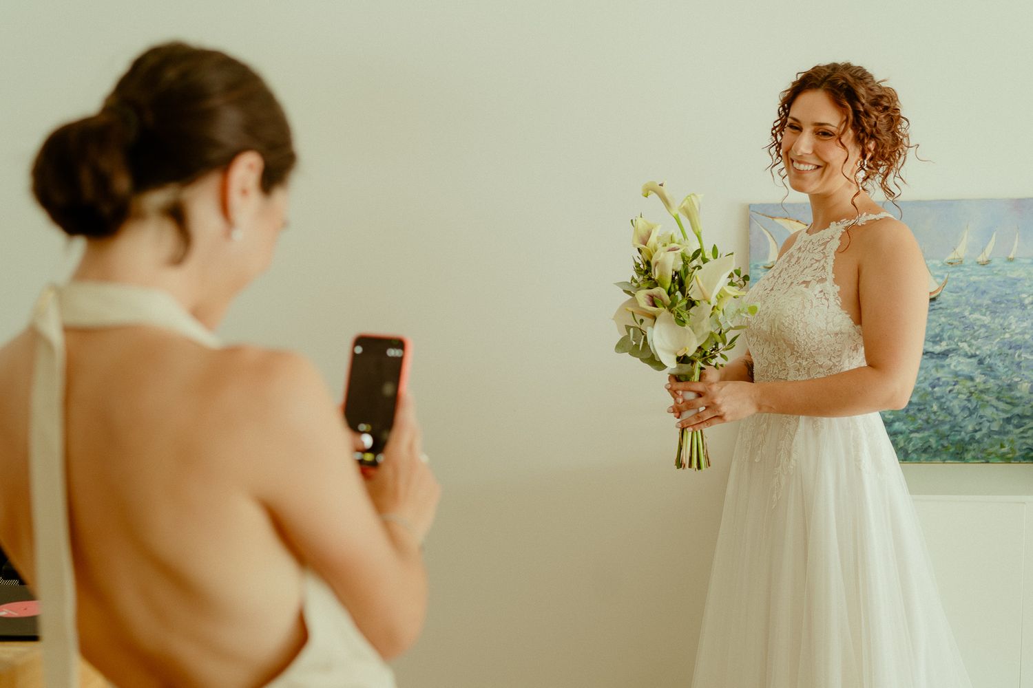 Bridesmaid photographs smiling bride in white gown holding bouquet of white flowers in bright indoor setting.