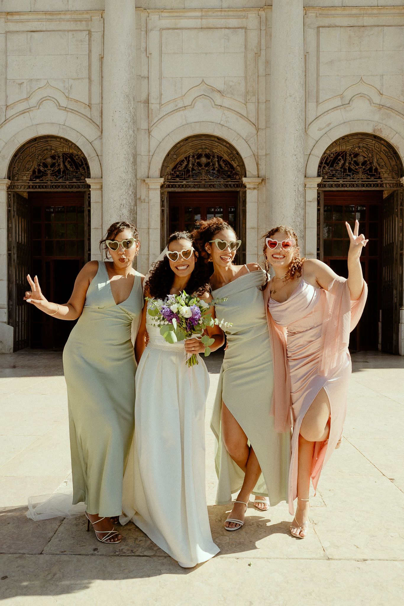 Bride and three bridesmaids in sage green dresses pose with peace signs and sunglasses outside a grand building.