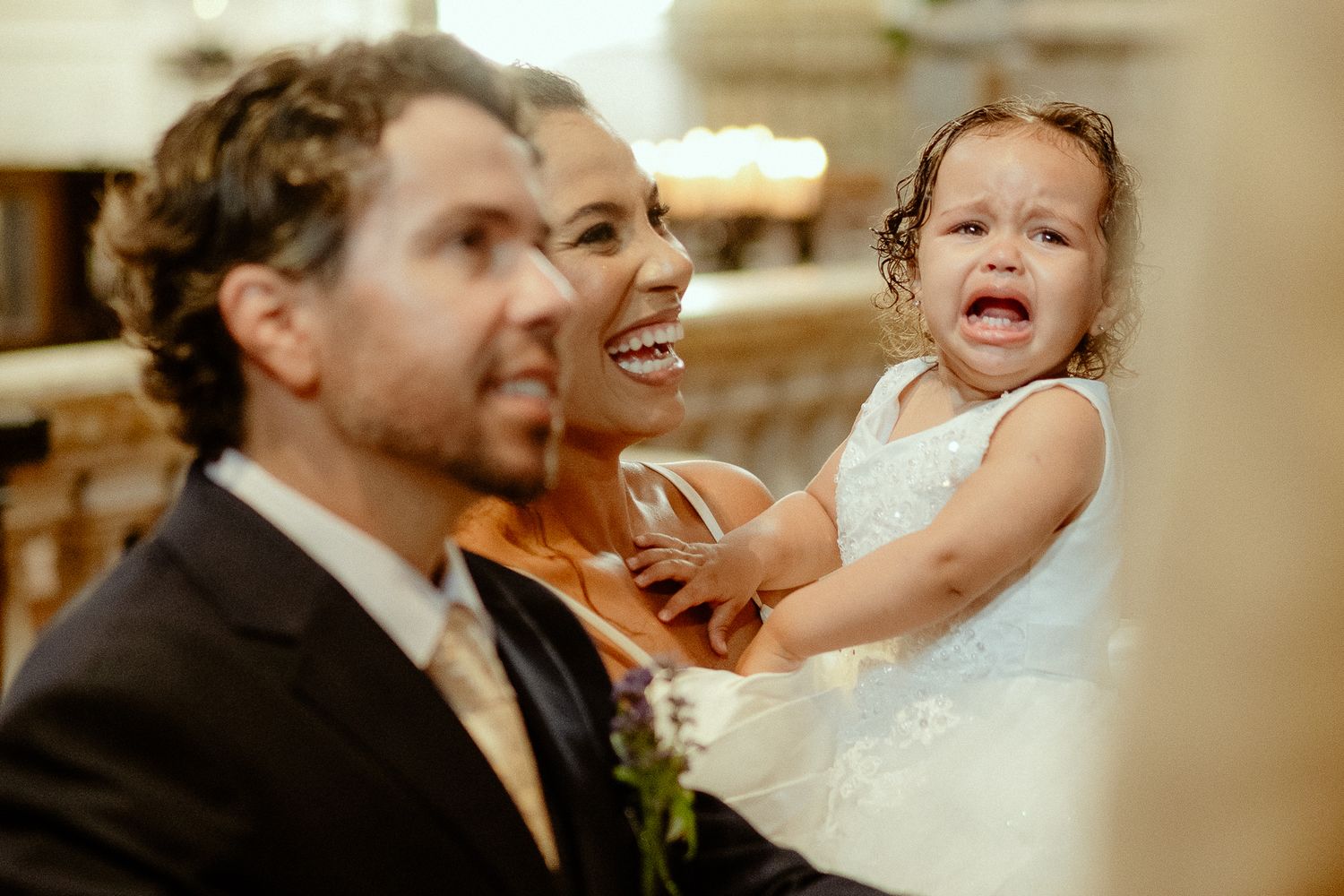 A bride laughing joyfully while holding a crying flower girl in a white dress during a wedding ceremony.