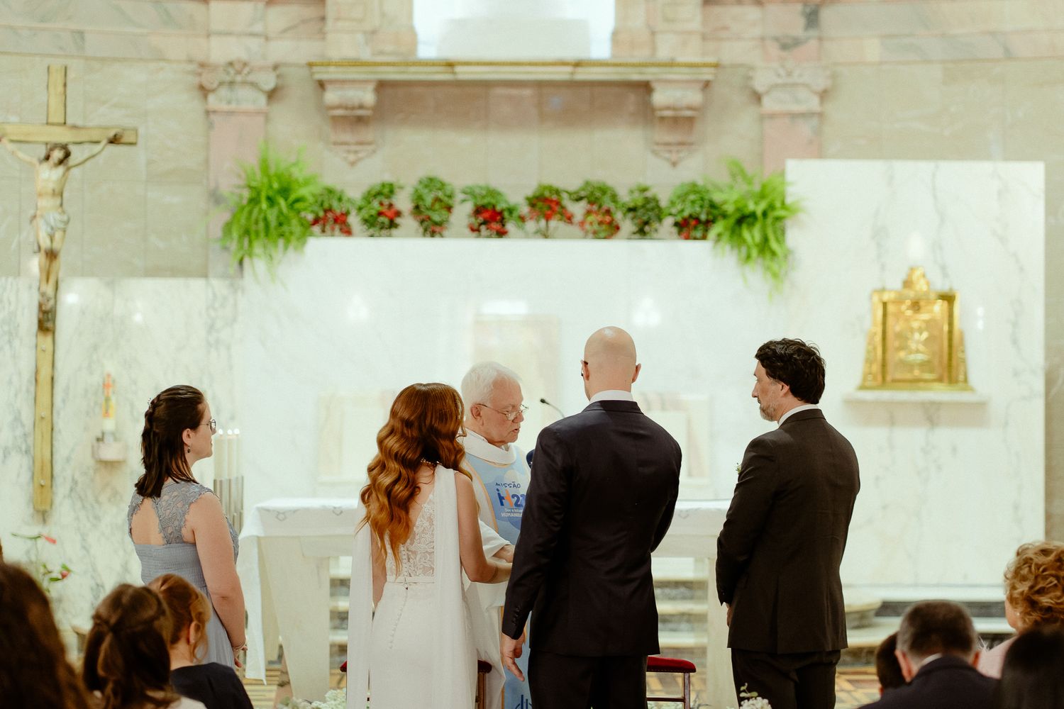 A wedding ceremony inside a church with a couple at the altar, guests seated, and floral greenery decorating the backdrop.