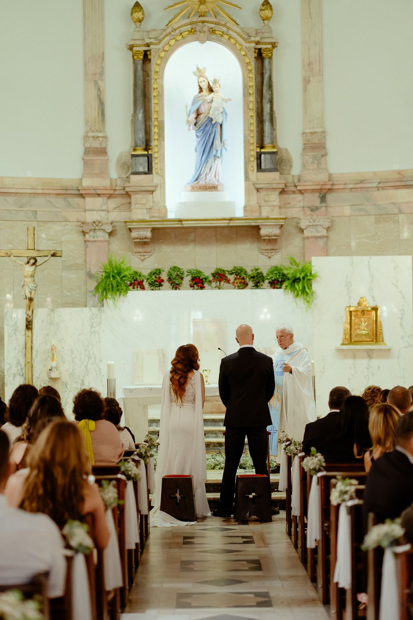 Couple exchanging vows at Catholic church altar with priest, guests seated in pews, Virgin Mary statue in background.