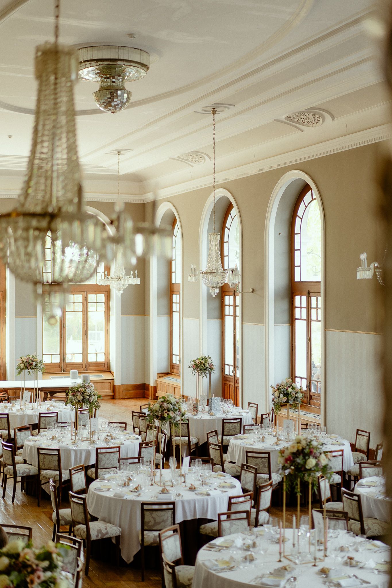 Elegant wedding reception hall with round tables, white linens, floral centerpieces, crystal chandeliers, and tall arched windows.