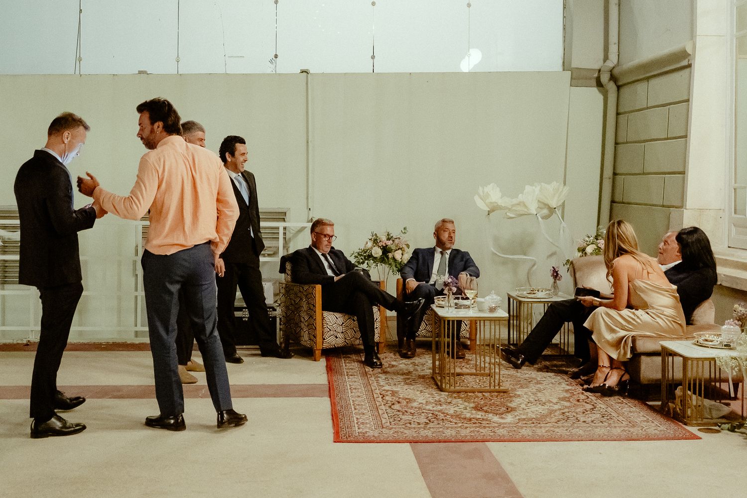 Elegantly dressed guests mingle and relax at an upscale indoor event with floral arrangements and gold furniture.