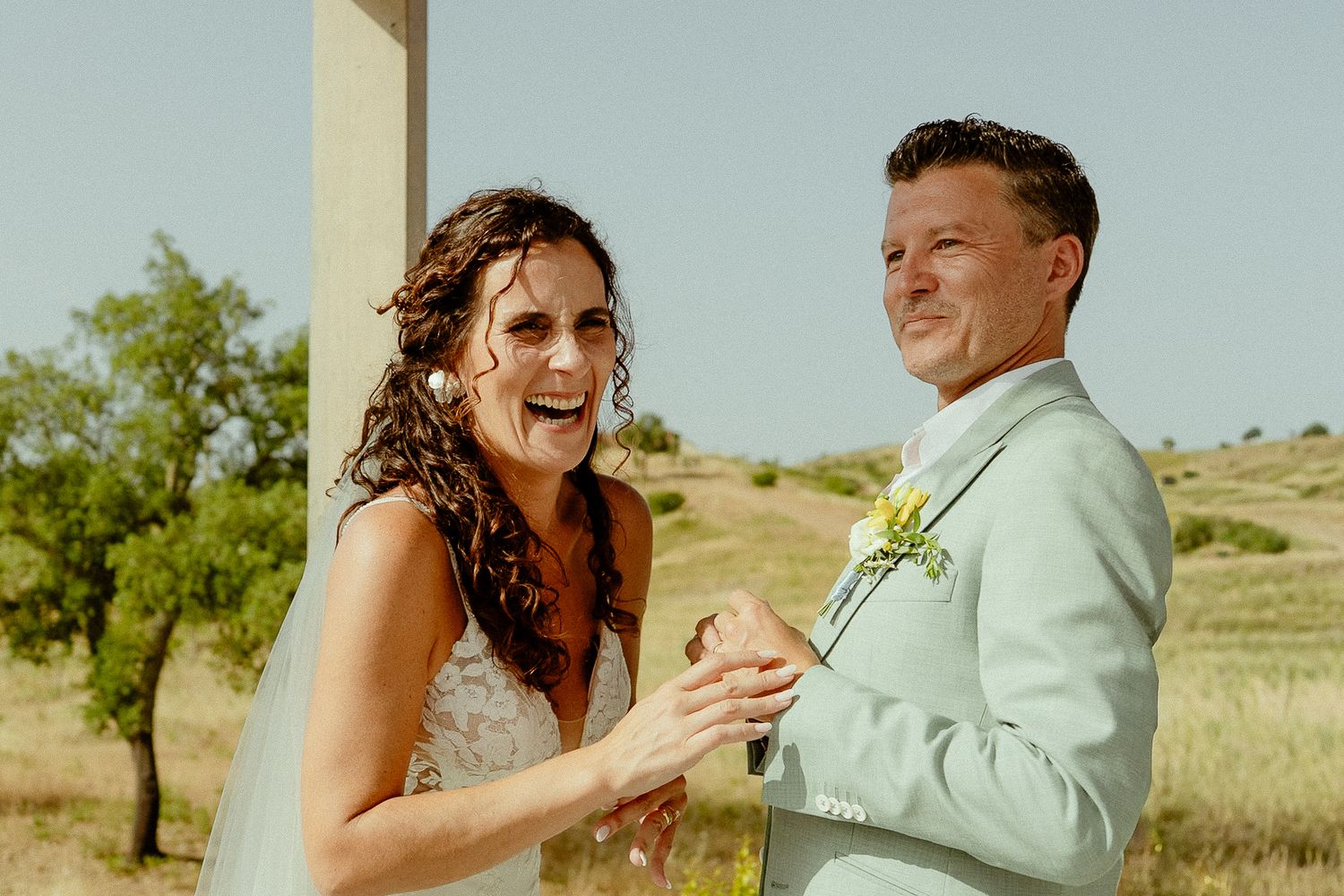 Joyful bride and groom laughing together outdoors on their wedding day in a sunny field.