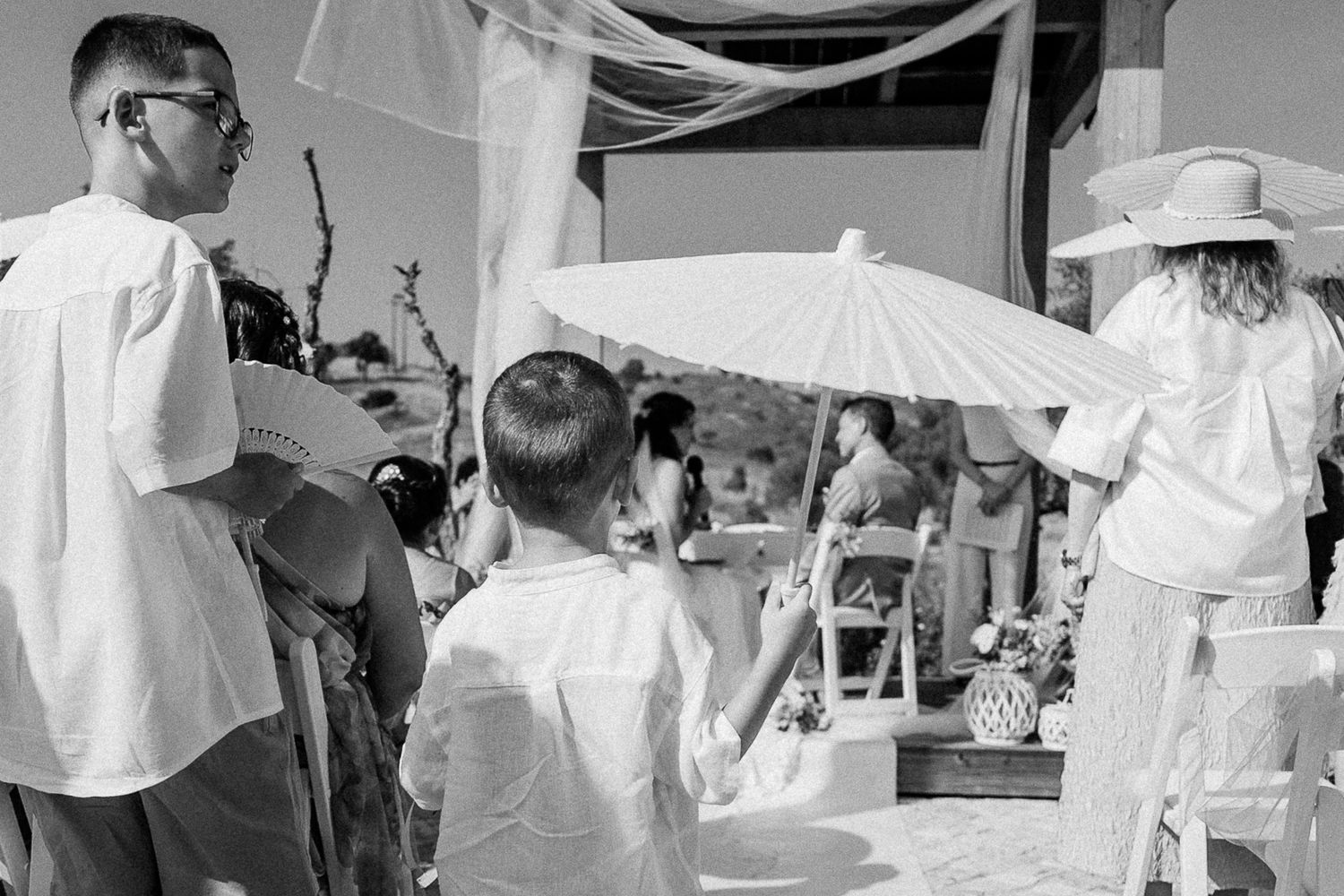 Black and white beach wedding ceremony with guests holding white parasols in elegant white attire.