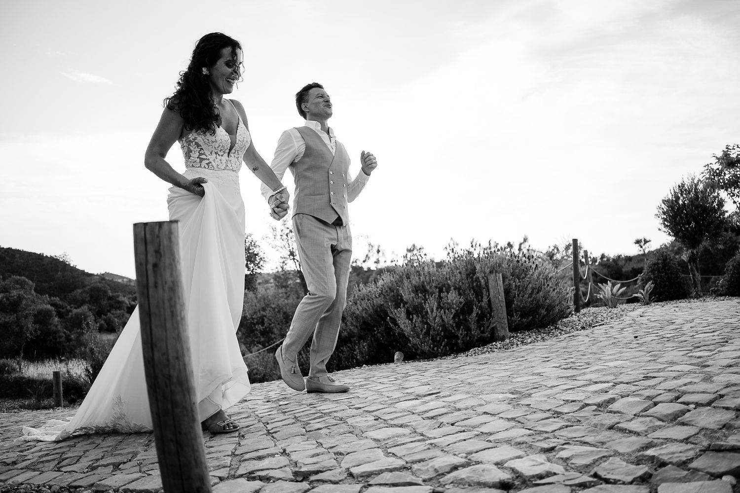 Black and white photo of a bride and groom holding hands on a cobblestone path, groom jumping joyfully.