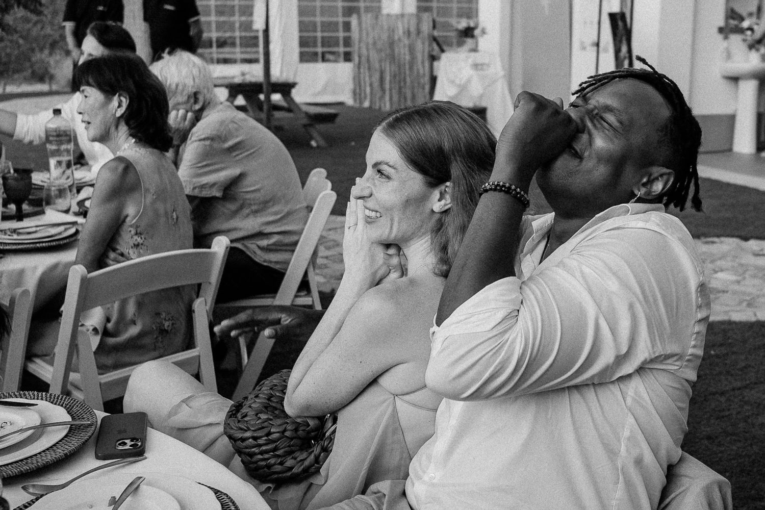 Black and white photo of a couple laughing and enjoying an outdoor wedding reception dinner.
