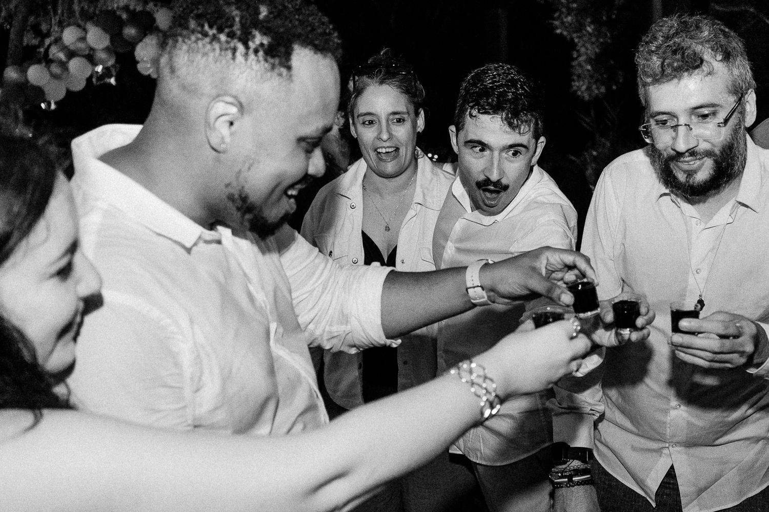 Black and white photo of a group of friends toasting with shot glasses at a wedding reception party.