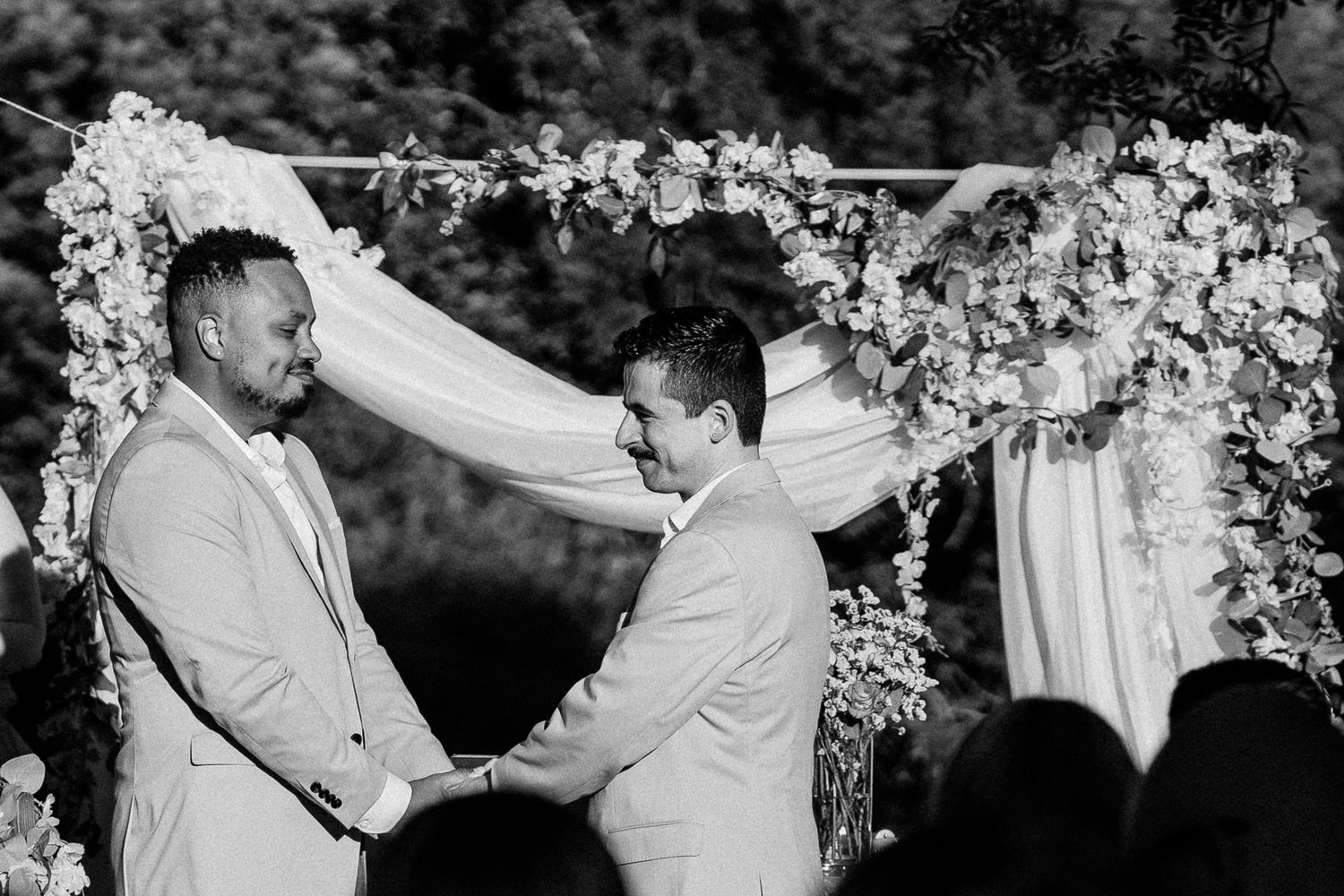 Two grooms exchange vows under a floral arch in a black and white outdoor wedding ceremony photo.