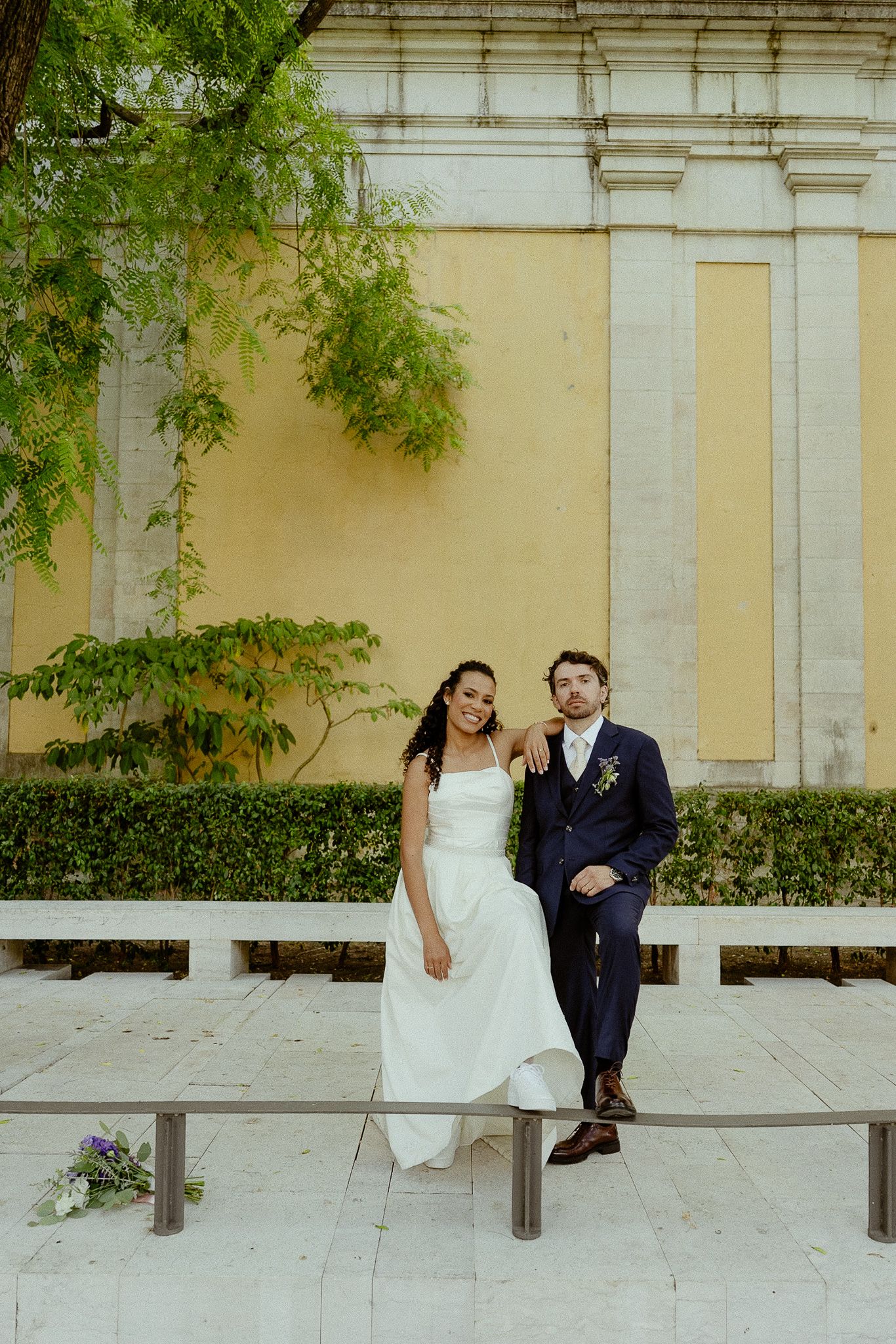 A bride and groom sit together on a bench outdoors, smiling, with a yellow wall and green trees behind them.