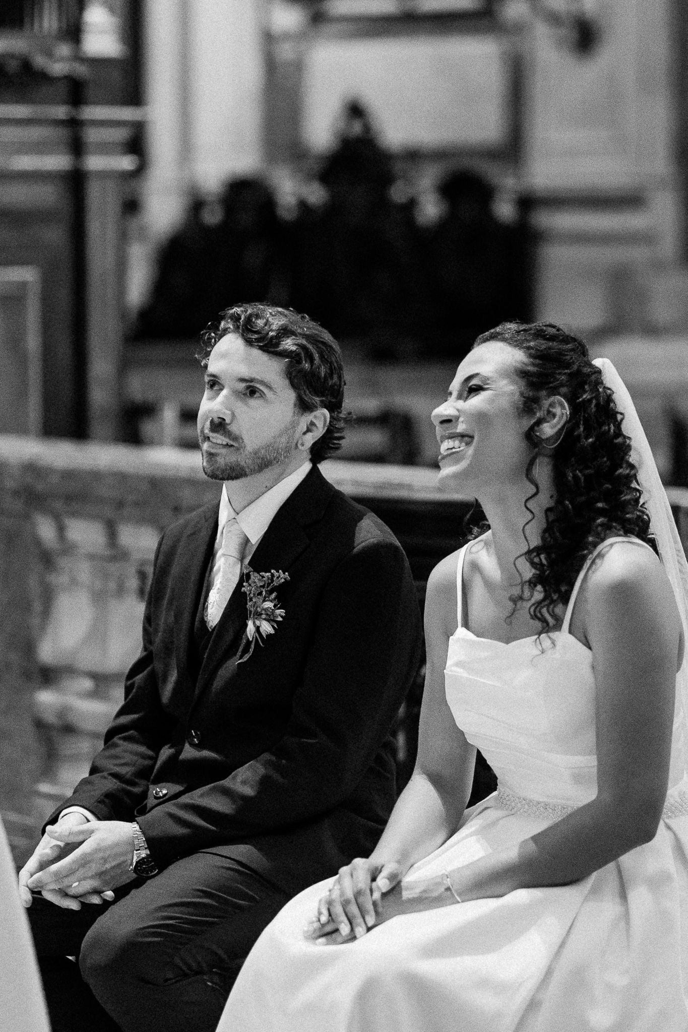 Black and white photo of a bride and groom sitting together during their wedding ceremony in an elegant venue.