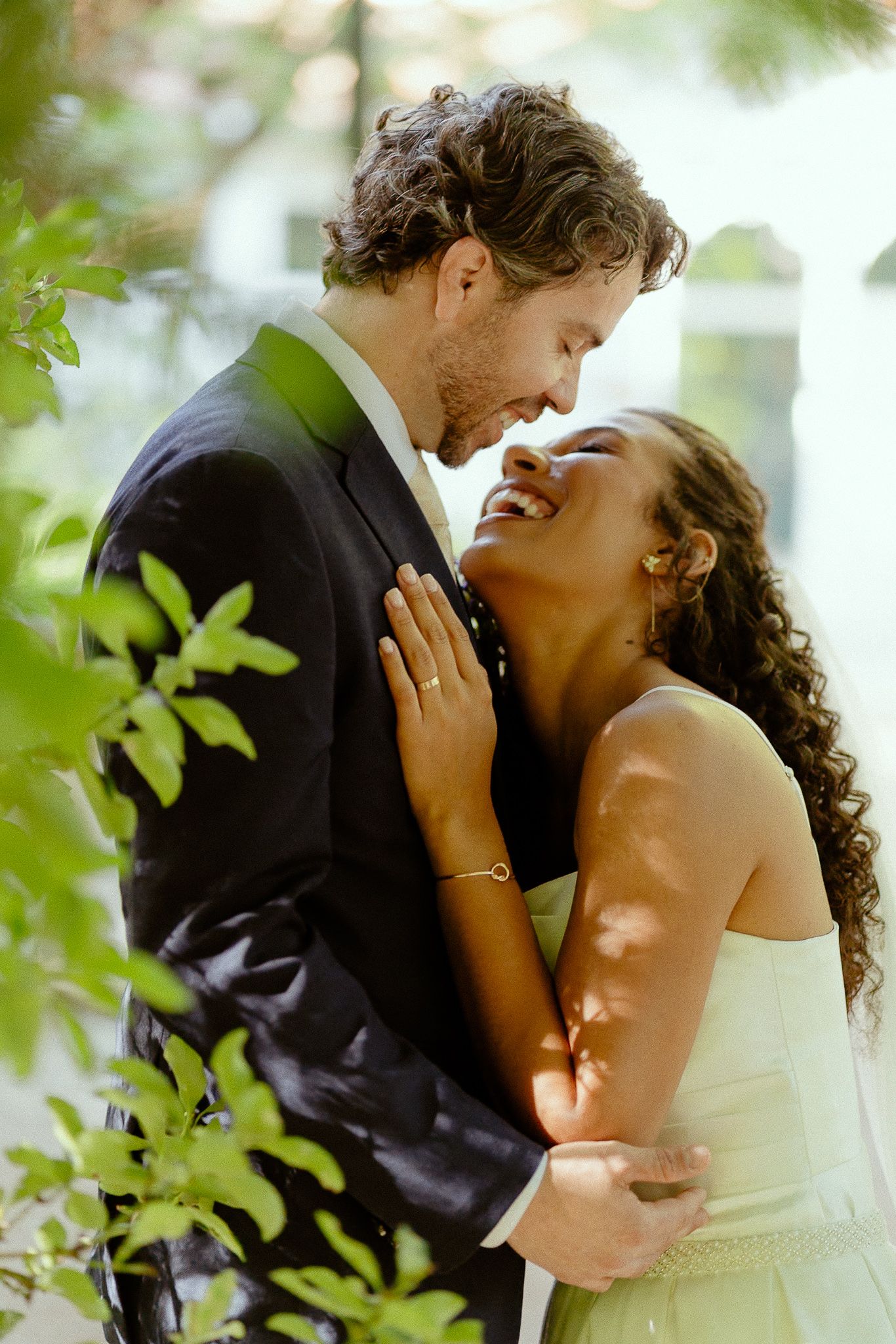 A joyful interracial couple in wedding attire embrace outdoors, the bride laughing as groom holds her close.