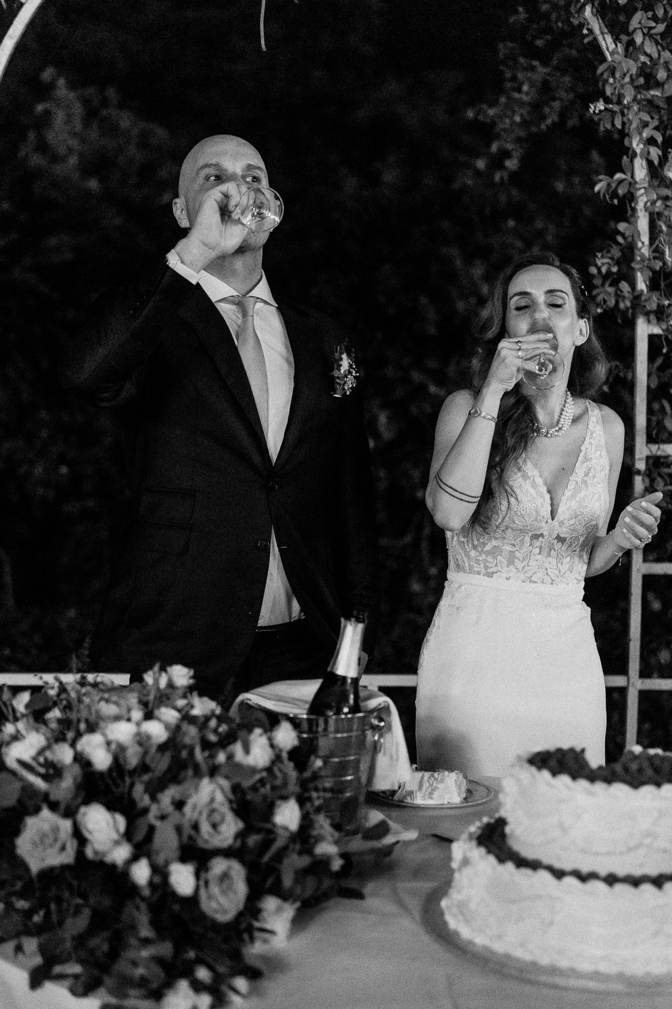 Black and white photo of bride and groom toasting with champagne beside wedding cake and floral arrangements.