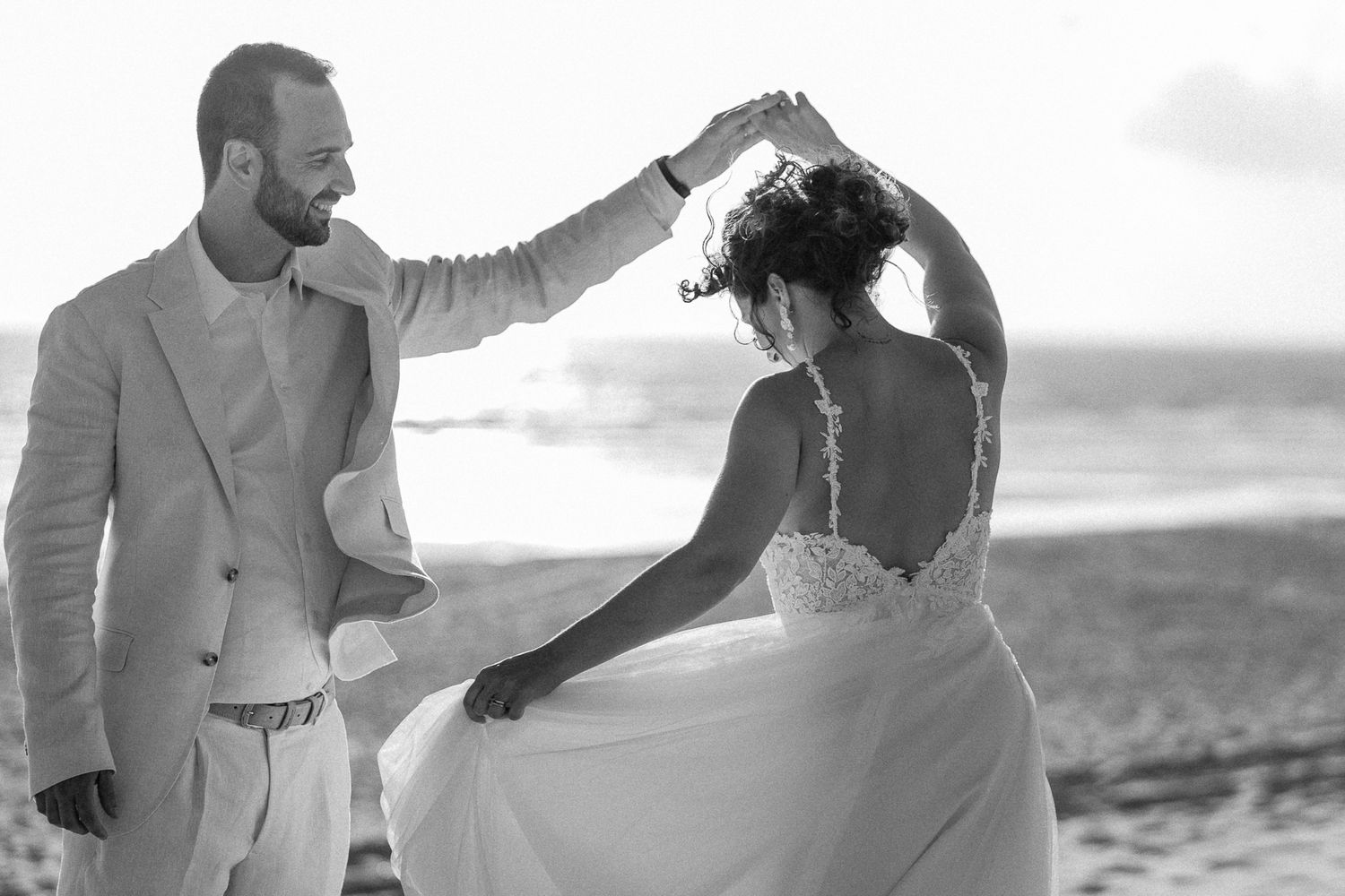 Black and white beach wedding photo of groom spinning bride in white lace gown by the ocean.