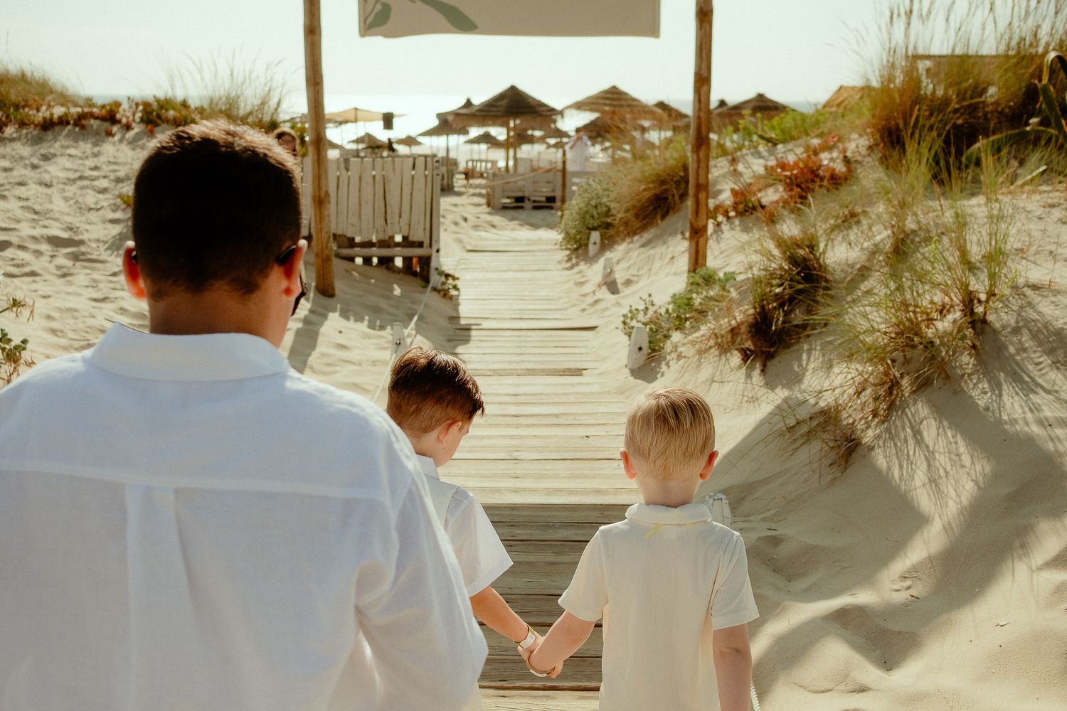 Father walks two young boys in white shirts down a wooden boardwalk path to a sandy beach.