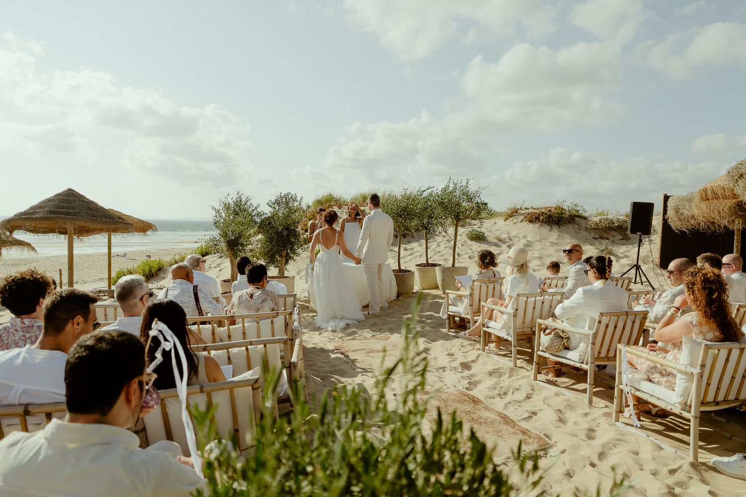 Romantic beach wedding ceremony with guests seated on chairs in the sand near the ocean on a sunny day.