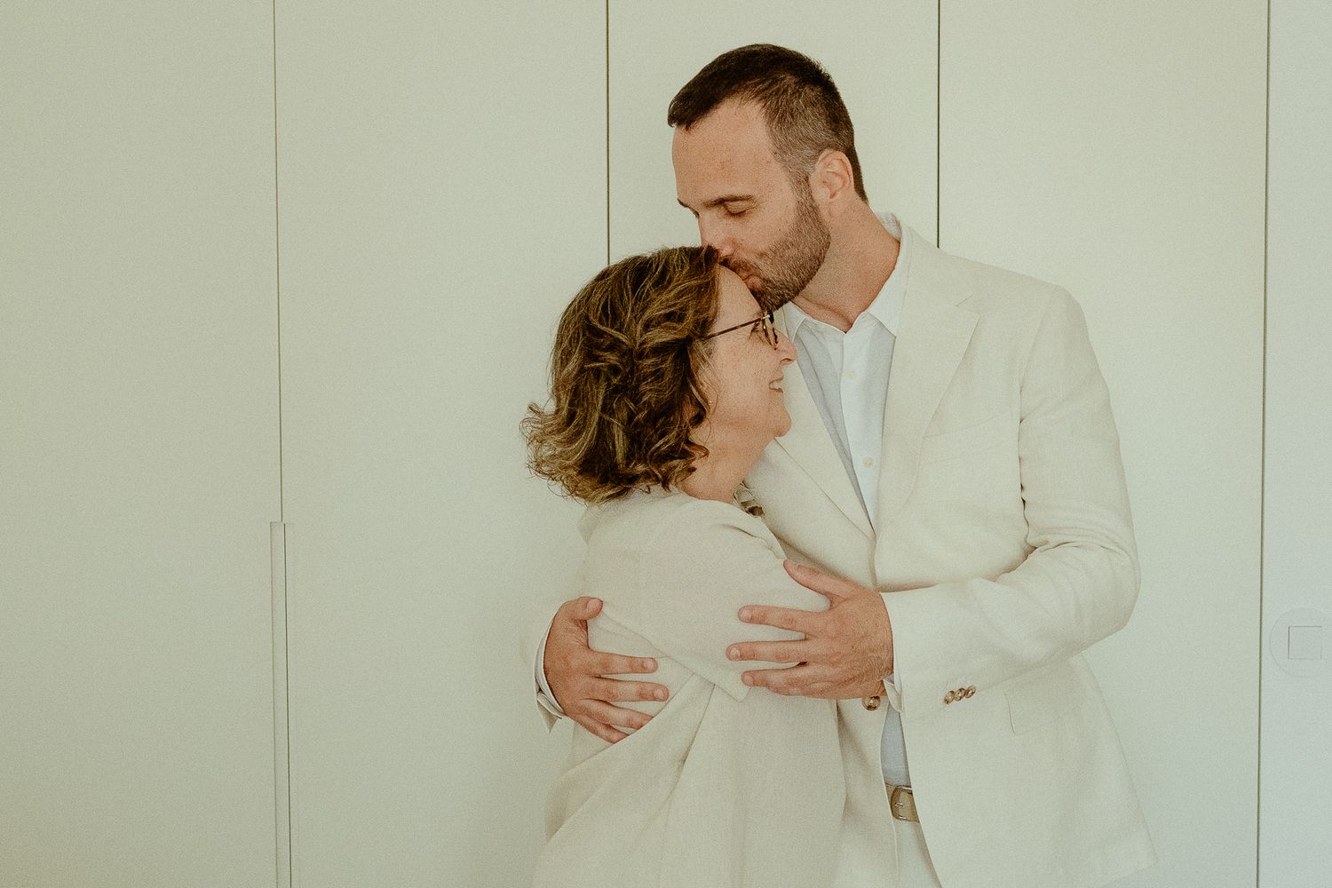 A man in a white suit kisses a pregnant woman's head while tenderly cradling her baby bump against a white wall.
