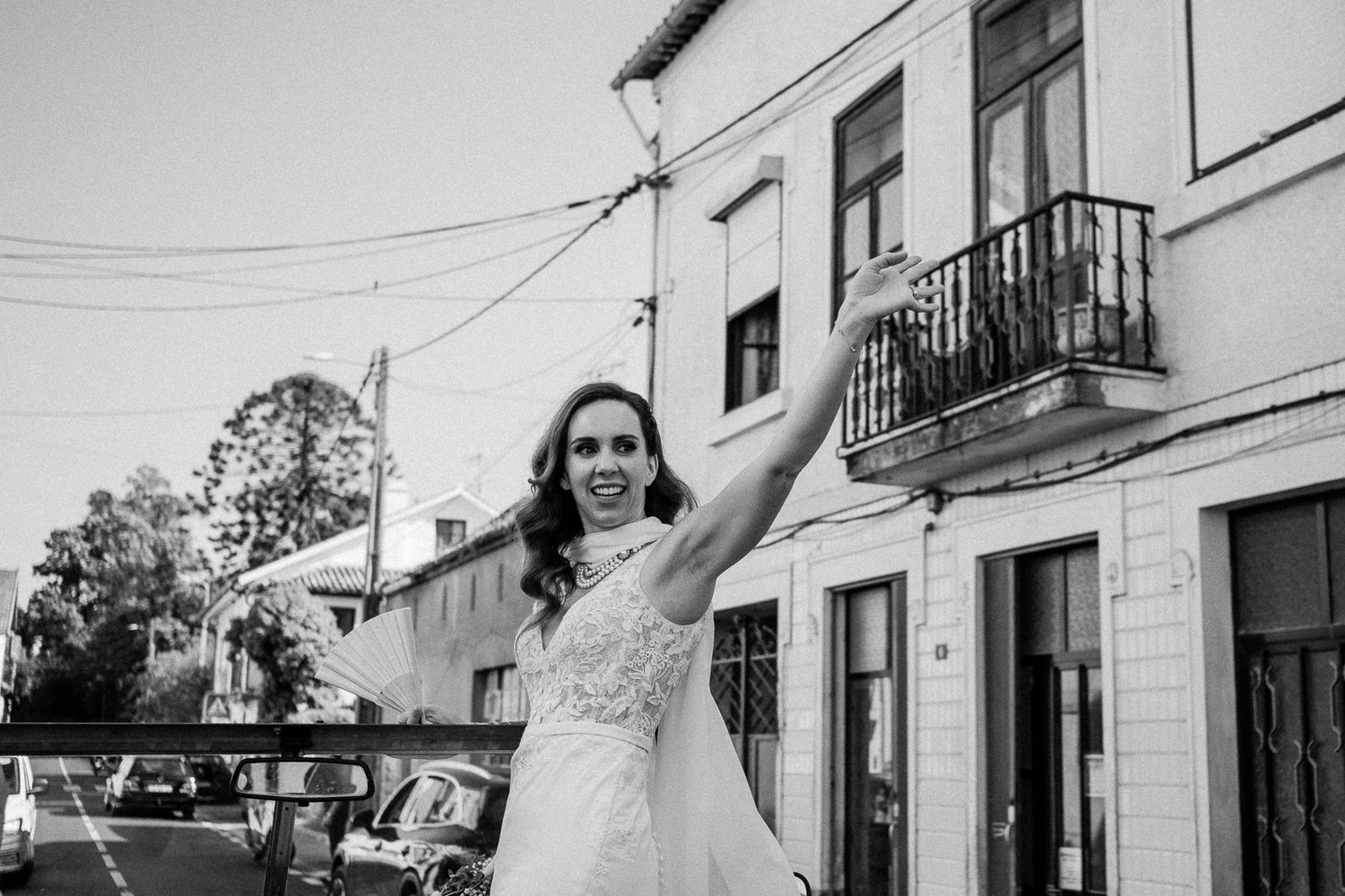Bride in white lace gown waves joyfully on a street, black and white wedding photo.