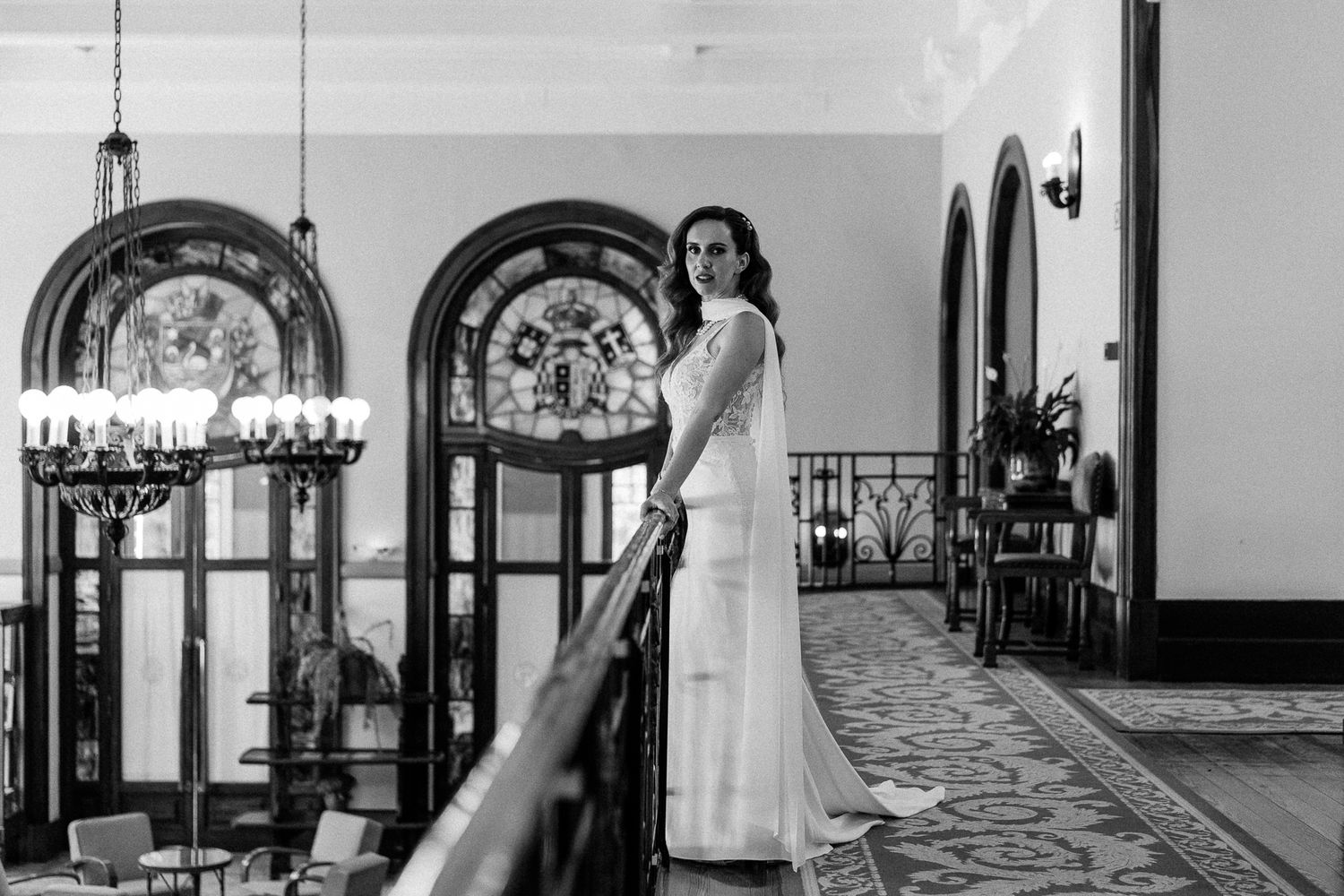 Bride in white gown poses on elegant staircase in black and white wedding photo with ornate decor.