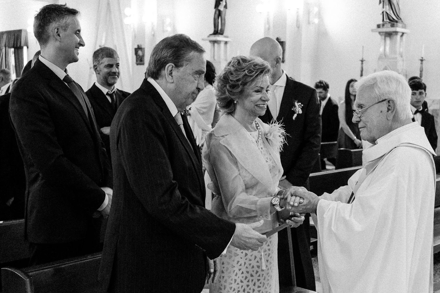 Black and white wedding ceremony photo of couple exchanging rings with priest in elegant church setting.