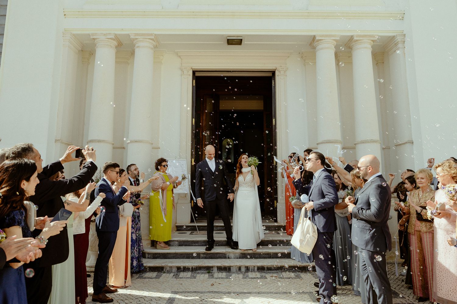 Newlywed couple exits a white church greeted by guests throwing confetti during a wedding ceremony.