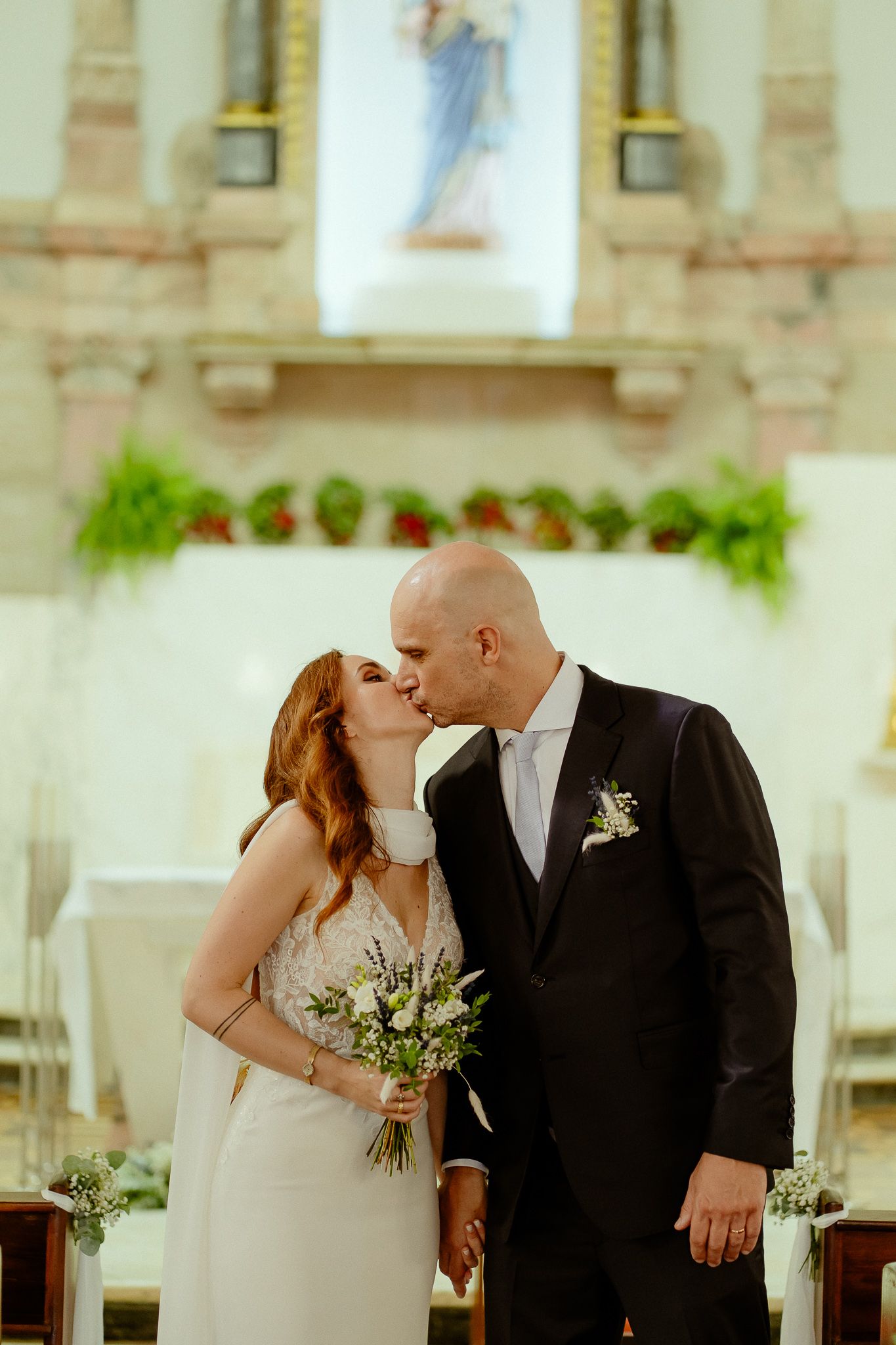 Bride and groom share a kiss at the altar of a decorated church, bride holding a white floral bouquet.