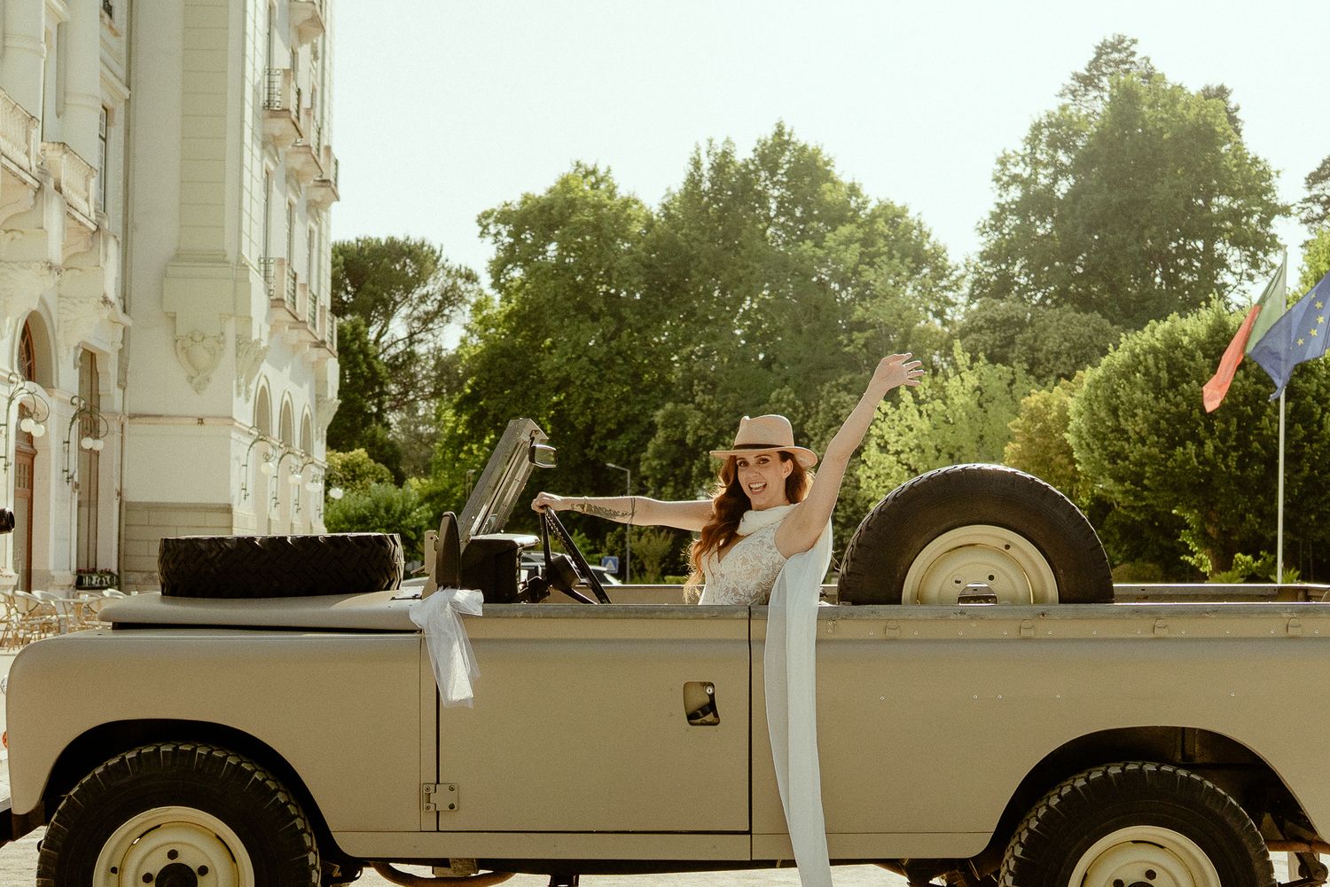 Woman in hat posing beside a vintage beige Land Rover convertible near a grand building surrounded by trees.