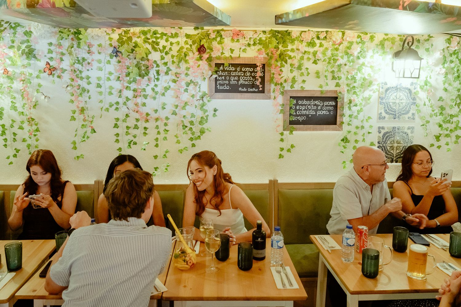 Guests dining at a cozy restaurant with green vine wall decor, wooden tables, and casual seating.
