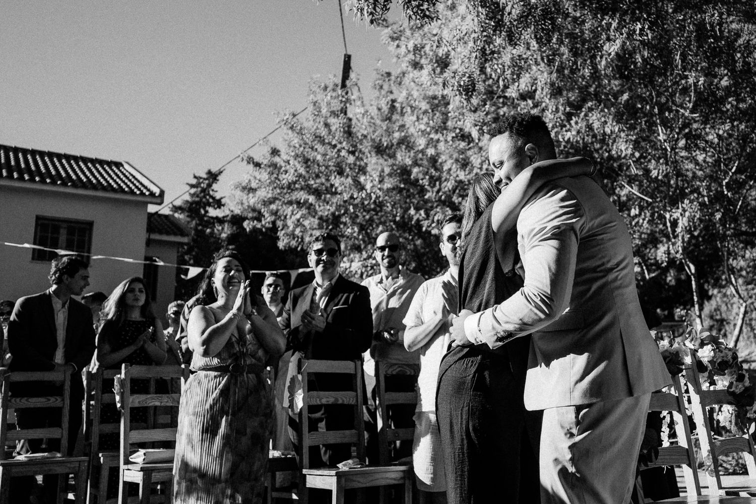 Black and white outdoor wedding ceremony with guests applauding as couple embraces near chairs and trees.