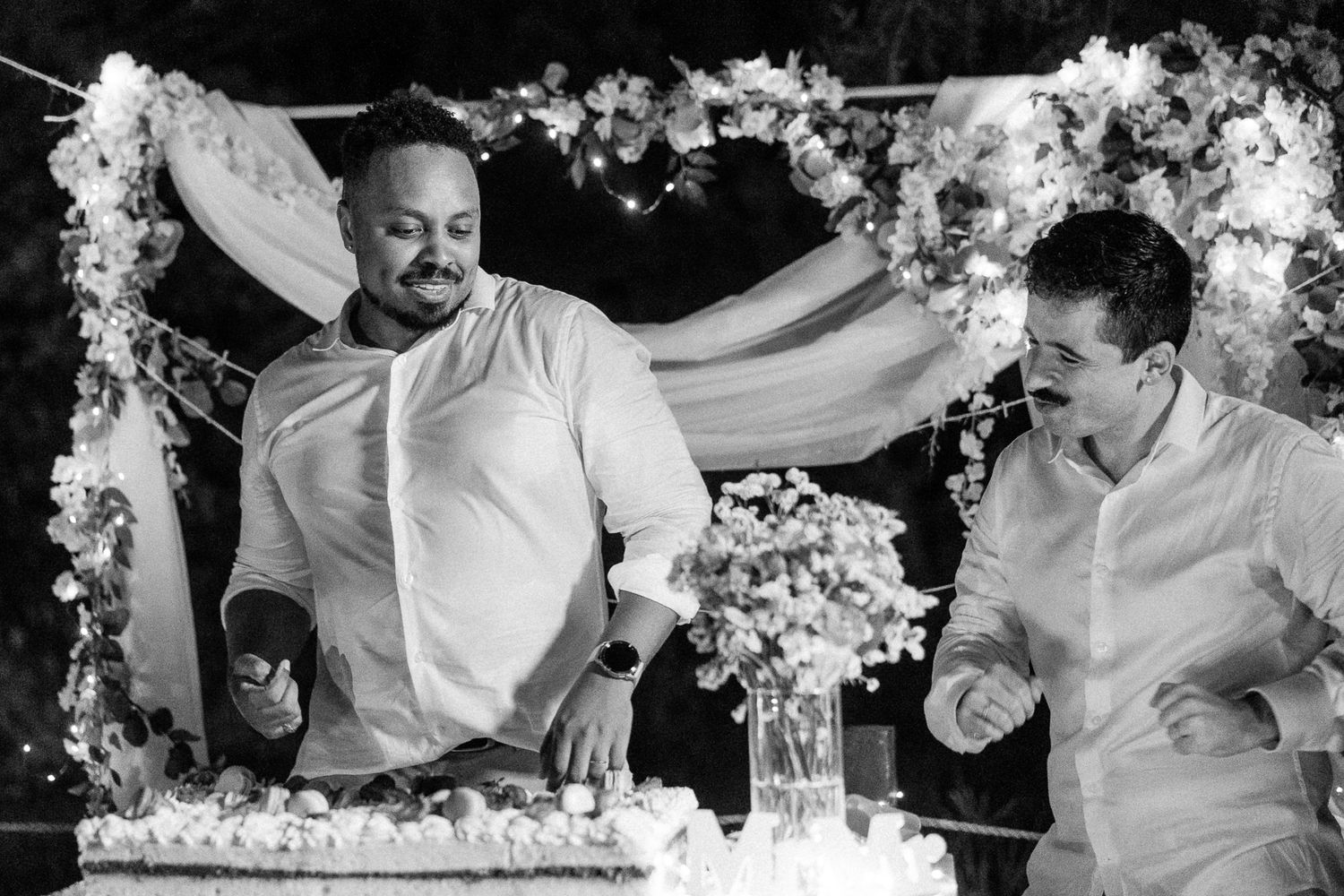 Two men in white shirts celebrate joyfully at a wedding cake cutting ceremony under floral arch decorations.