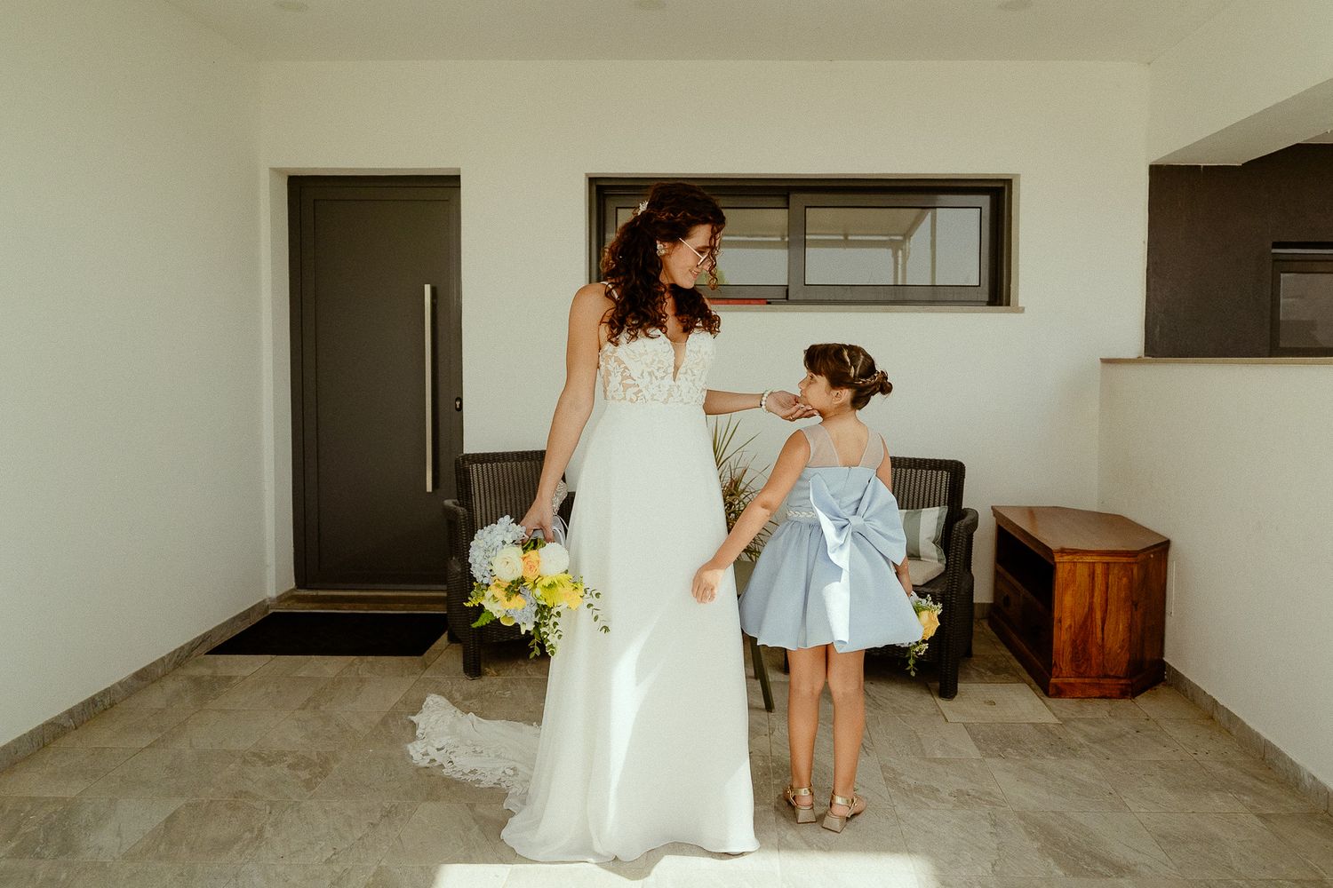 Bride in white gown holds yellow bouquet while interacting with flower girl in blue dress indoors.
