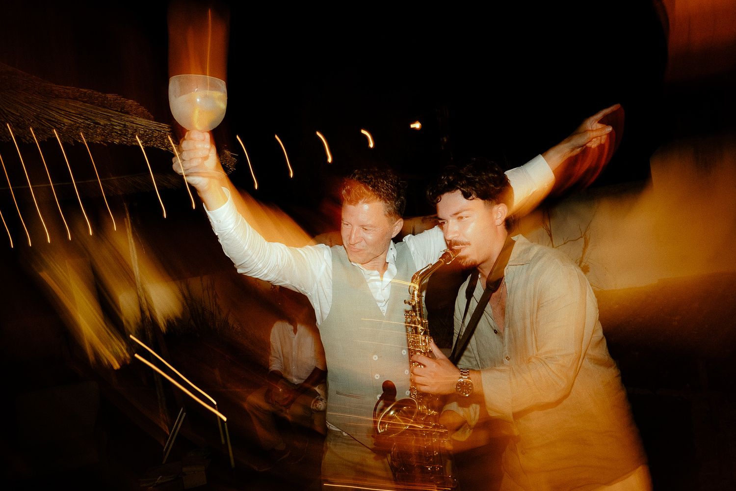 Two men celebrate at night, one raising a glass and the other playing saxophone, captured in a dynamic blurred motion shot.