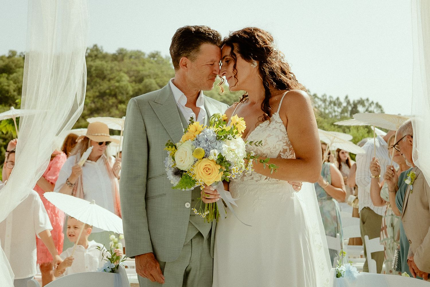 Bride and groom share a romantic moment at outdoor wedding ceremony, bride holding yellow and blue bouquet.
