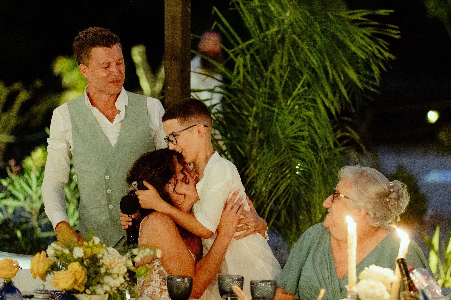 Couple embraces at outdoor wedding reception dinner table with candles and floral arrangements at night.