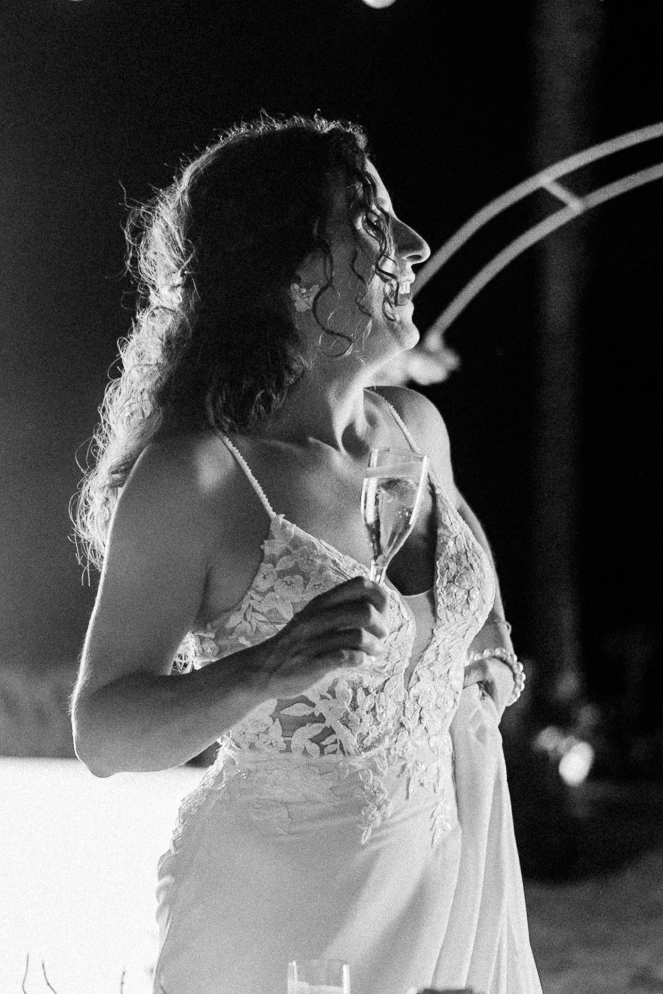 Black and white photo of a smiling bride in lace dress holding champagne glass at wedding reception.