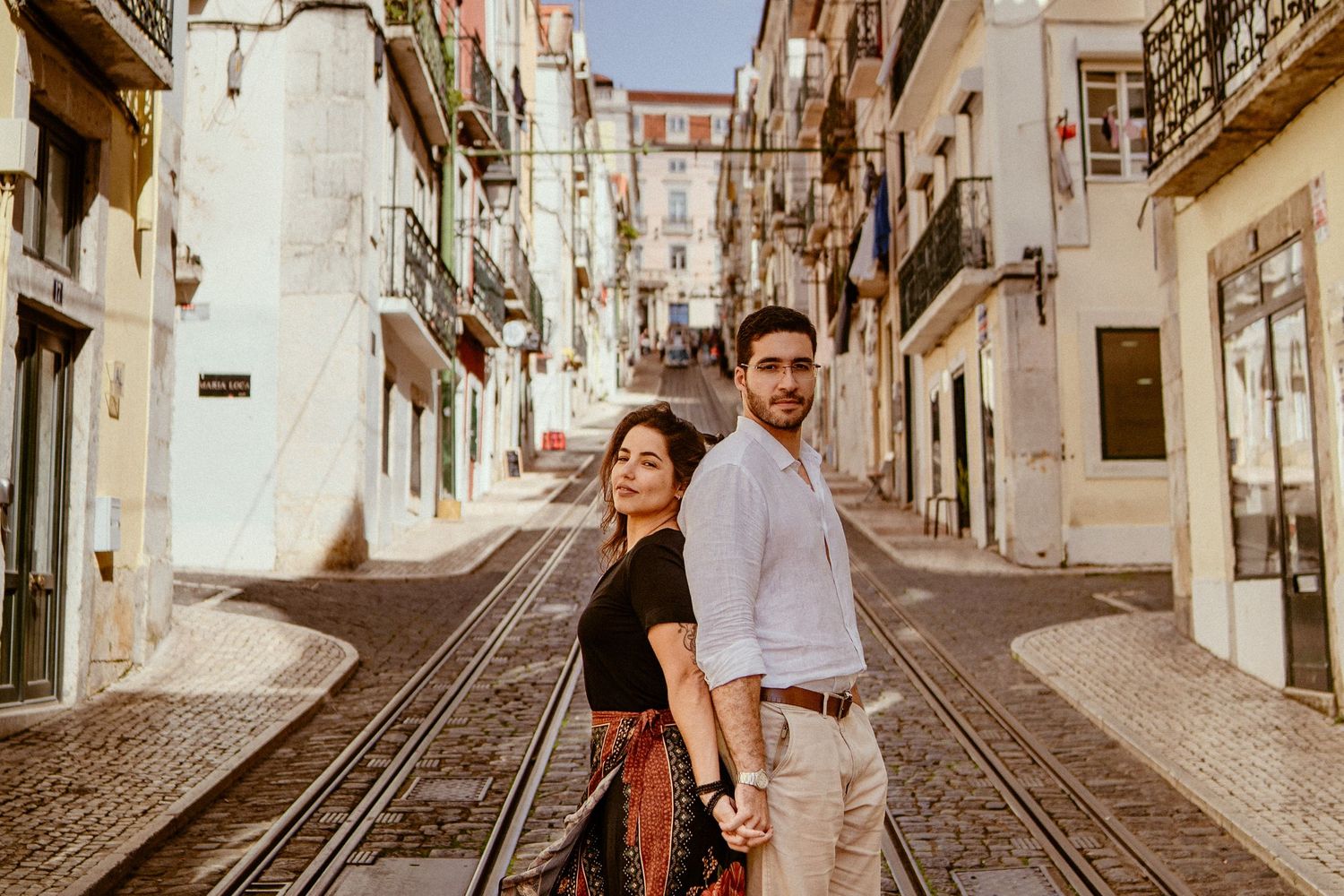 Two people pose back-to-back on historic Lisbon street with tram tracks running down cobblestone path.