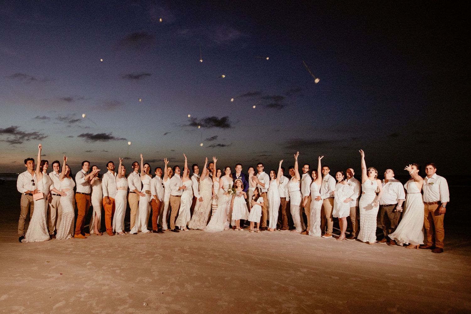 Large wedding party in all-white attire poses together on beach at dusk with starlit sky above.