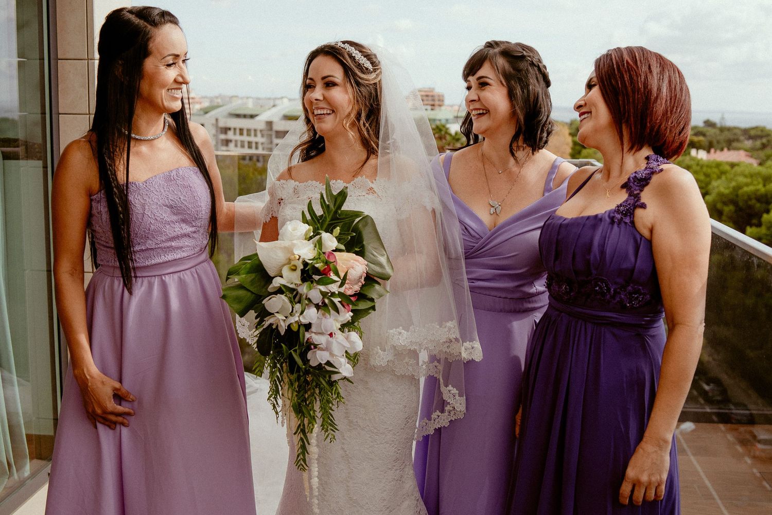Bridesmaids in purple dresses share joyful moments on a balcony with city views in the background.