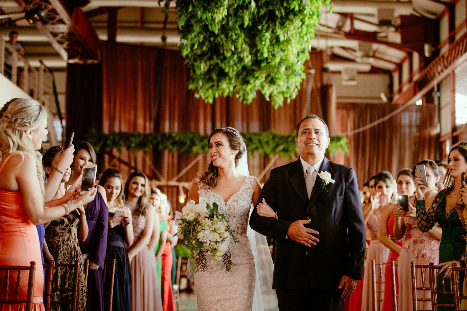 Wedding guests dancing joyfully at an indoor reception with string lights and greenery hanging from wooden beams.