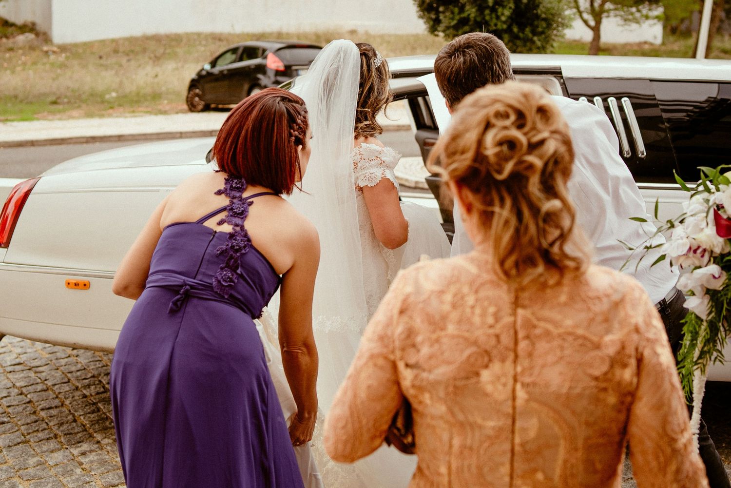 Wedding party helping bride into white limousine on sunny day.